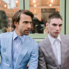 Dwóch mężczyzn w garniturach w kratę. Jeden w błękitnym, drugi w brązowym. Eleganckie klapy w marynarce podkreślają ich styl.